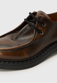 Chaussure en cuir brun avec une finition lisse, bout rond, lacets noirs et semelle en caoutchouc texturée présentant un design en zigzag.