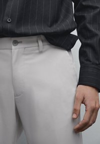 Helle graue Hose mit glatter Textur und taillierter Passform, kombiniert mit einem dunkel gestreiften Hemd mit Knöpfen. Verfügt über eine dezente Vordertasche.