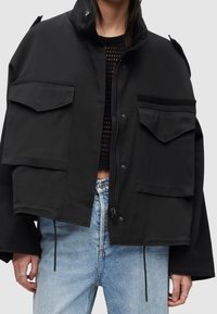 Zwarte cropped jas met hoge kraag, ritssluiting, twee grote voorzakken en gestructureerde accenten; gecombineerd met lichtblauwe spijkerbroek.