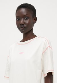 T-shirt a maniche corte di colore crema con cuciture rosa, caratterizzato dal logo "HUGO" sul petto. Tessuto morbido con una vestibilità rilassata.