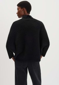 Schwarzer, übergroßer Strickpullover mit strukturiertem Finish und hohem Kragen. Lässig geschnittene Ärmel mit subtiler Formgebung, kombiniert mit dunklen Jeans.