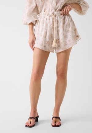 Vrouw draagt beige korte broek met schelpenprint, taillekoord, beige top en donkere sandalen met hak, staand tegen een witte achtergrond.