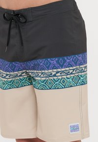 Svømmebukser med en svart toppseksjon, mønstret midtseksjon i lilla og teal, og beige bunn. Har snorstramning og merkeetikett.