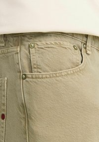 Khaki corduroy bukser med en struktureret overflade, der har metalbeslag og en lille rød accent nær lommen.
