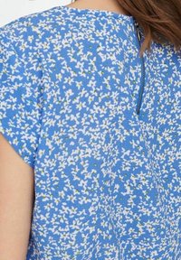 Tessuto blu con piccole stampe di fiori bianchi e gialli, che mostra i dettagli delle maniche corte e della zip sul retro di un indumento.