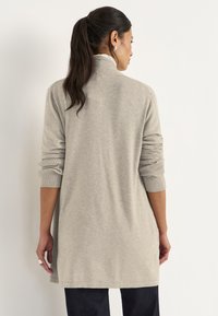 Vrouw met een donkere ponytail die een lichtgrijze lange cardigan en donkere broek draagt, staand met haar gezicht naar een witte muur.
