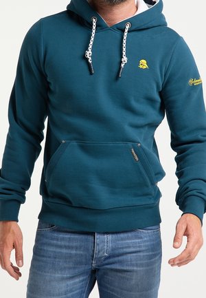 Homme portant un sweat à capuche turquoise avec cordons blancs, logos jaunes sur la poitrine et la manche, et une poche avant, associé à un jean bleu.