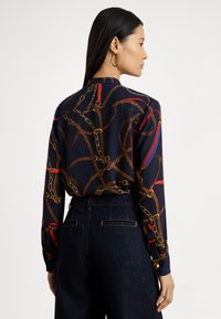 Kobieta z długimi ciemnymi włosami, nosząca złote kolczyki koła, granatową bluzkę z nadrukiem łańcuchów w kolorach brązowym i czerwonym oraz ciemne jeansy z wysokim stanem, zwrócona bokiem.