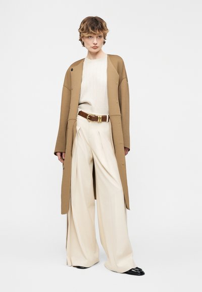 Άτομο που φοράει μπεζ φαρδιά παντελόνια, λευκό πουλόβερ, καφέ ζώνη, μακρύ παλτό καμηλό και μαύρα παπούτσια, στέκεται μπροστά σε λευκό φόντο.