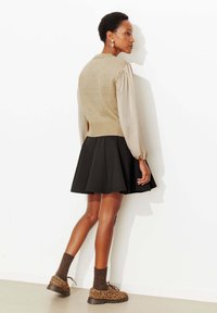 Pull tricot beige clair avec manches longues bouffantes, associé à une jupe évasée noire et des chaussures à imprimé léopard marron, debout contre un mur blanc.