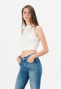 Bílý pruhovaný crop top s vysokým výstřihem a bez rukávů, kombinovaný s modrými džínami s vysokým pasem, které mají přední kapsy a zapínání na knoflík.