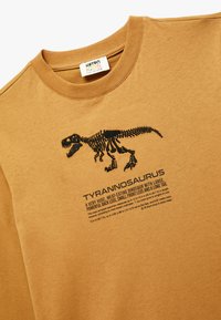 Ruskea puuvillapaita, jossa on musta Tyrannosaurus-skeletontulostus ja kuvaileva teksti alla. Pyöreä pääntie, pehmeä textura, lyhyet hihat.