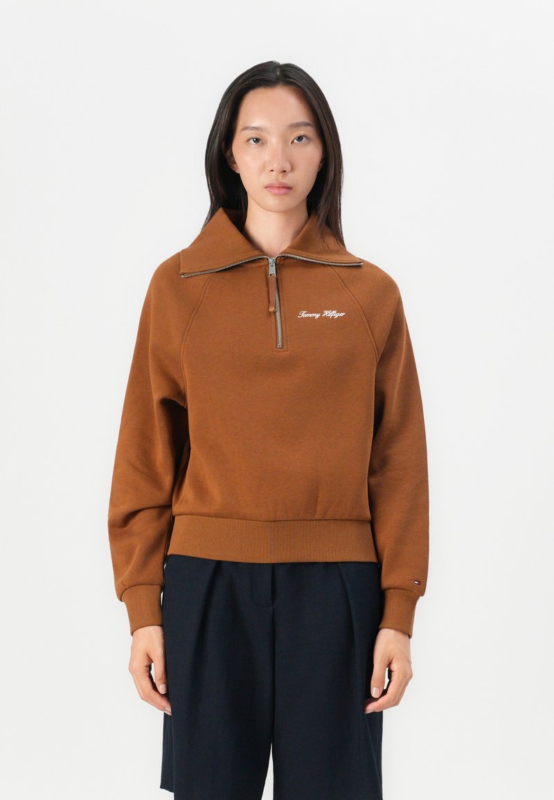 Brown zip-neck pullover sweatshirt with ribbed cuffs and hem. Features logo embroidery on the chest and a structured collar design.