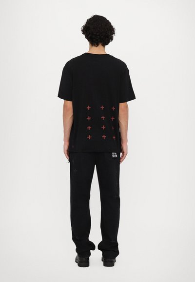 T-shirt noire avec un motif de croix rouge au dos, associée à un pantalon noir. Coupe carrée, manches courtes et texture en coton.