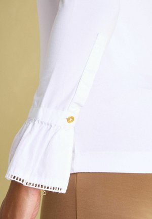 White blouse with billowy sleeves. Features a gold button detail and intricate lace trim at the cuff. Smooth texture, fitted body.