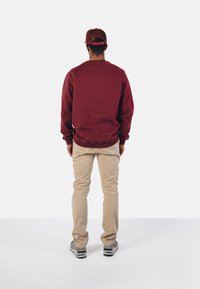 Maroon sweatshirt dengan cuff dan hem berbahan ribbed, dipadukan dengan celana beige terang dan sneakers abu-abu. Dilihat dari belakang dengan topi yang serasi.