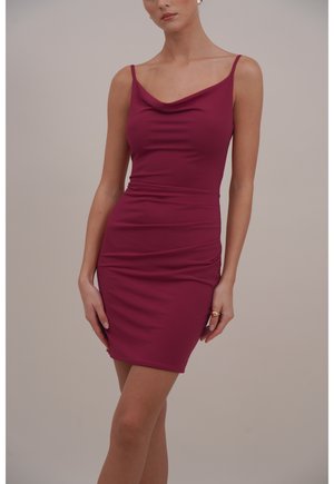 Kobieta ubrana w dopasowaną, bezrękawnikową, do kolan sukienkę w ciemnoróżowym kolorze z cienkimi ramiączkami i drapowanym dekoltem, stojąca na tle jednolitego tła.