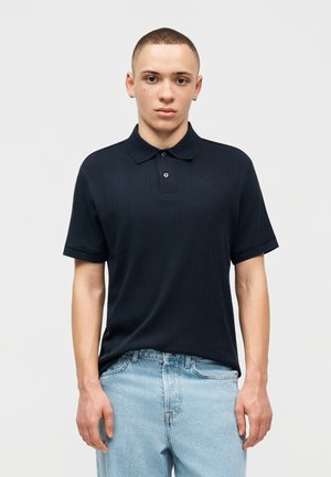 Νέος άνδρας με κοντά μαλλιά, φορεί ένα μαύρο πόλο με κοντά μανίκια και ανοιχτόχρωμα τζιν, στέκεται μπροστά σε ένα απλό λευκό φόντο.