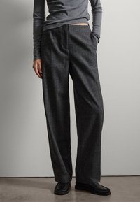 Pantalons larges gris foncé en tissu texturé, avec des poches latérales et un devant plat. Portés avec un haut gris ajusté et des chaussures noires.