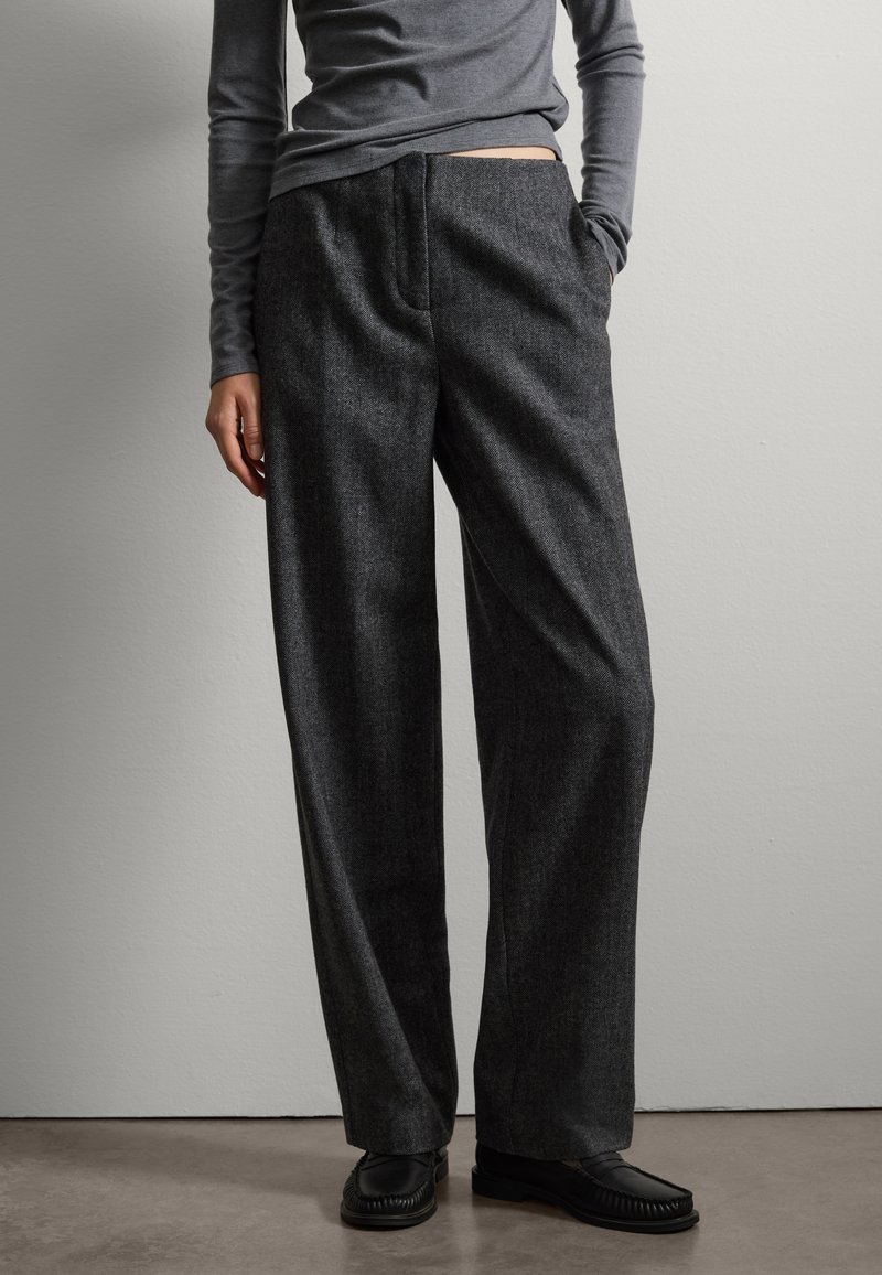Pantalons larges gris foncé en tissu texturé, avec des poches latérales et un devant plat. Portés avec un haut gris ajusté et des chaussures noires.