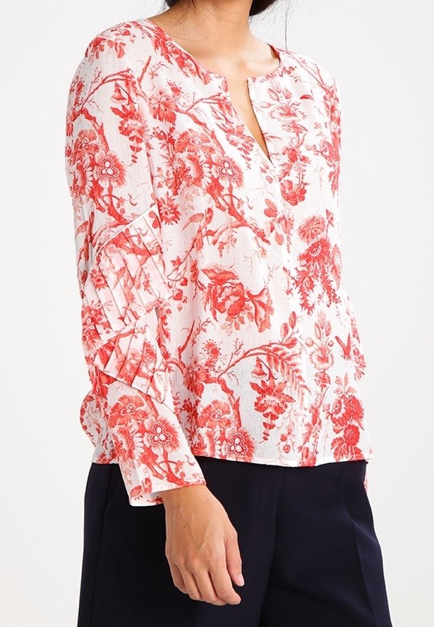 Blusa bianca con stampa floreale rossa, caratterizzata da scollatura a V e maniche lunghe con dettaglio arricciato. Tessuto leggero e texture.