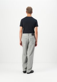 Mann med kort blondt hår iført en svart t-skjorte, lysegrå jeans og svarte sko som står vendt mot en hvit vegg.