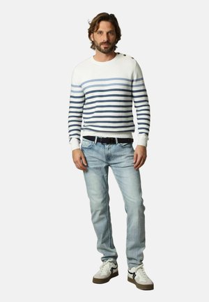 Homme debout portant un pull blanc à rayures bleues, un jean bleu clair, une ceinture noire et des baskets blanches sur fond gris clair.
