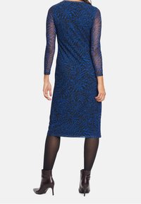 Žena oblečená v modro-černých vzorovaných šatech ke kolenům s průhlednými dlouhými rukávy, černými punčochami a tmavě hnědými kotníkovými botami, stojící zády.