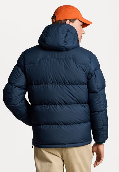 Giubbotto imbottito blu navy con cappuccio, caratterizzato da sezioni trapuntate, una texture liscia e tasche laterali con zip. Indossato con un berretto di colore arancione vivace.