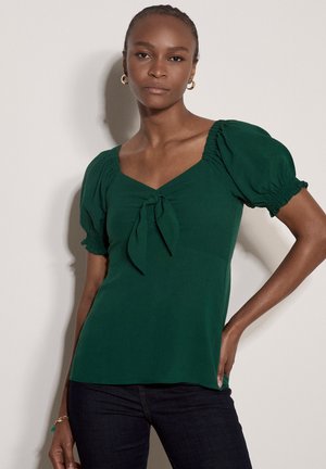 Kobieta z warkoczami, nosząca złote kolczyki-koła i zieloną bluzkę z bufkami i wiązaniem z przodu, pozująca na neutralnym tle.