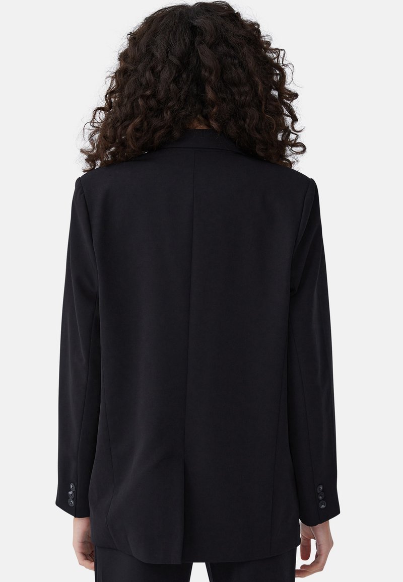 Osoba z długimi kręconymi włosami, ubrana w czarną marynarkę z guzikami na rękawach, pokazana od tyłu na białym tle.