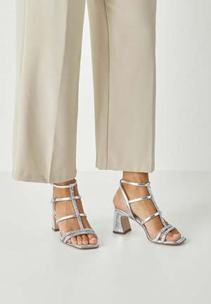 Srebrne sandały na obcasie z wieloma paskami i ozdobami z kamieniami. Obcas ma kształt blokowy, zapewniając stabilność i styl.