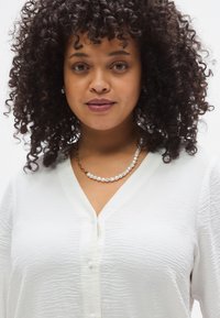 Λευκό, υφασμάτινο πουκάμισο με V-λαιμόκοψη και κουμπιά, σε συνδυασμό με κολιέ πολλαπλών σειρών από πέρλες που εμφανίζει μείγμα από πέρλες και μεταλλικές λεπτομέρειες.