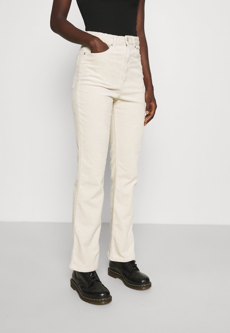 Personne portant un pantalon en velours côtelé écru taille haute, un haut noir et des bottes noires à lacets, debout avec une main dans la poche.