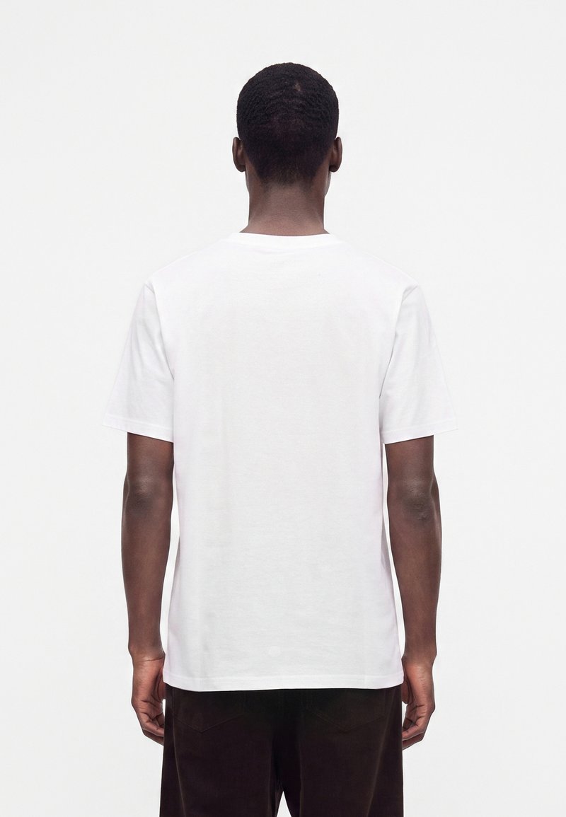 Mann mit kurzen dunklen Haaren, der ein schlichtes weißes T-Shirt mit kurzen Ärmeln und dunkle Hosen trägt, steht mit dem Rücken zur Kamera vor einem weißen Hintergrund.