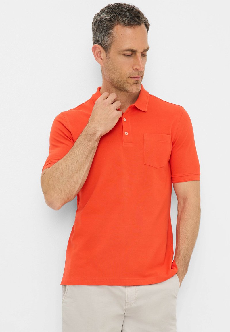 T-shirt polo orange avec un col et des manches courtes. Présente une poche sur la poitrine gauche et un tissu lisse. Porté avec un short beige clair.