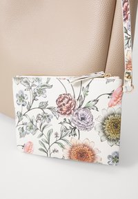 Petit porte-poignet blanc avec imprimé floral multicolore, fermeture éclair dorée et sac en cuir beige texturé en arrière-plan.