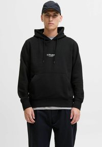 Schwarzer Pullover mit Kapuze und Kordelzug, vordere Kängurutasche und weißem Logo auf der Brust. Getragen mit dunklen Hosen und einer schwarzen Kappe.