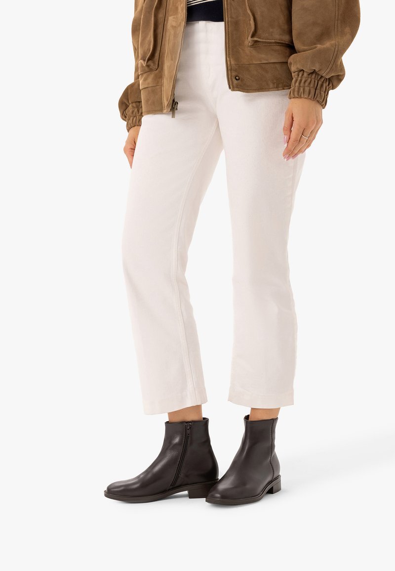 Personne portant un pantalon blanc court, des bottines marron foncé avec des fermetures éclair latérales, et une veste en suède marron avec des poignets élastiques.