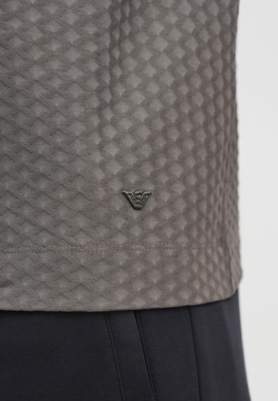 Γκρι υφασμάτινη υφή με διακοσ�μητικό μοτίβο διαμαντιού, που διαθέτει ένα μικρό μαύρο λογότυπο στο κάτω μέρος. Το ένδυμα συνδυάζεται με σκούρο παντελόνι.