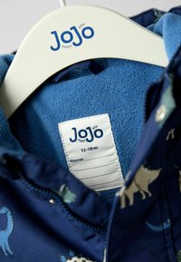 Námornícky modrá detská bunda so vzorom dinosaurov, mäkké fleecové podšívka, zipsové zapínanie a štítok s označením veľkosti 12-18 mesiacov. Viditeľné značkovanie JoJo.