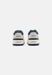 Zapatillas Lacoste que combinan ante beige y malla azul marino, con una suela y detalles en el talón blancos. El número "1933" está grabado en la parte trasera.