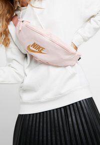 Růžový nylonový bum bag s výrazným zlatým logem Nike, vybavený zipem a nastavitelným popruhem, nošený přes bílou mikinu a černou plisovanou sukni.