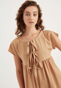 Beige strukturiertes Kleid mit kurzen Ärmeln, einem runden Ausschnitt und einer vorderen Bindeschleife. Fließendes Design mit gerafftem Taillenbereich.