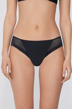 Culotte bikini noire à taille moyenne avec panneaux latéraux en mesh transparent portée par une personne debout sur un fond uni.