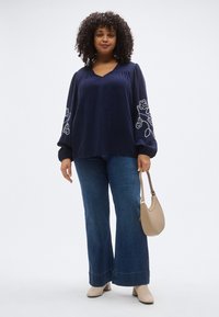 Blouse bleu marine à manches longues avec broderie florale blanche sur les manches, associée à un jean bleu évasé et un sac à main beige.