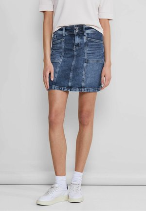 Dunkelblaue Jeans-Minirock mit figurbetontem Design, Fronttaschen und Knopfverschluss. Der Saum endet auf Höhe des Oberschenkels. Kombiniert mit weißen Sneakers.