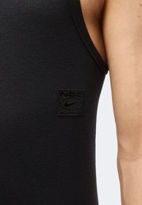 Gros plan d'une personne portant un débardeur noir en maille côtelée avec un petit patch noir du logo Nike sur la poitrine.