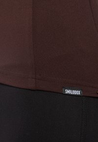 Haut de sport marron à texture lisse, avec une étiquette noire portant l'inscription "SMILODOX." Associé à un legging noir, avec un détail de couture.
