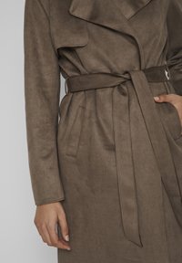 Trench-coat en daim marron avec un large col, une taille ceinturée, deux poches latérales et une texture douce et brossée.